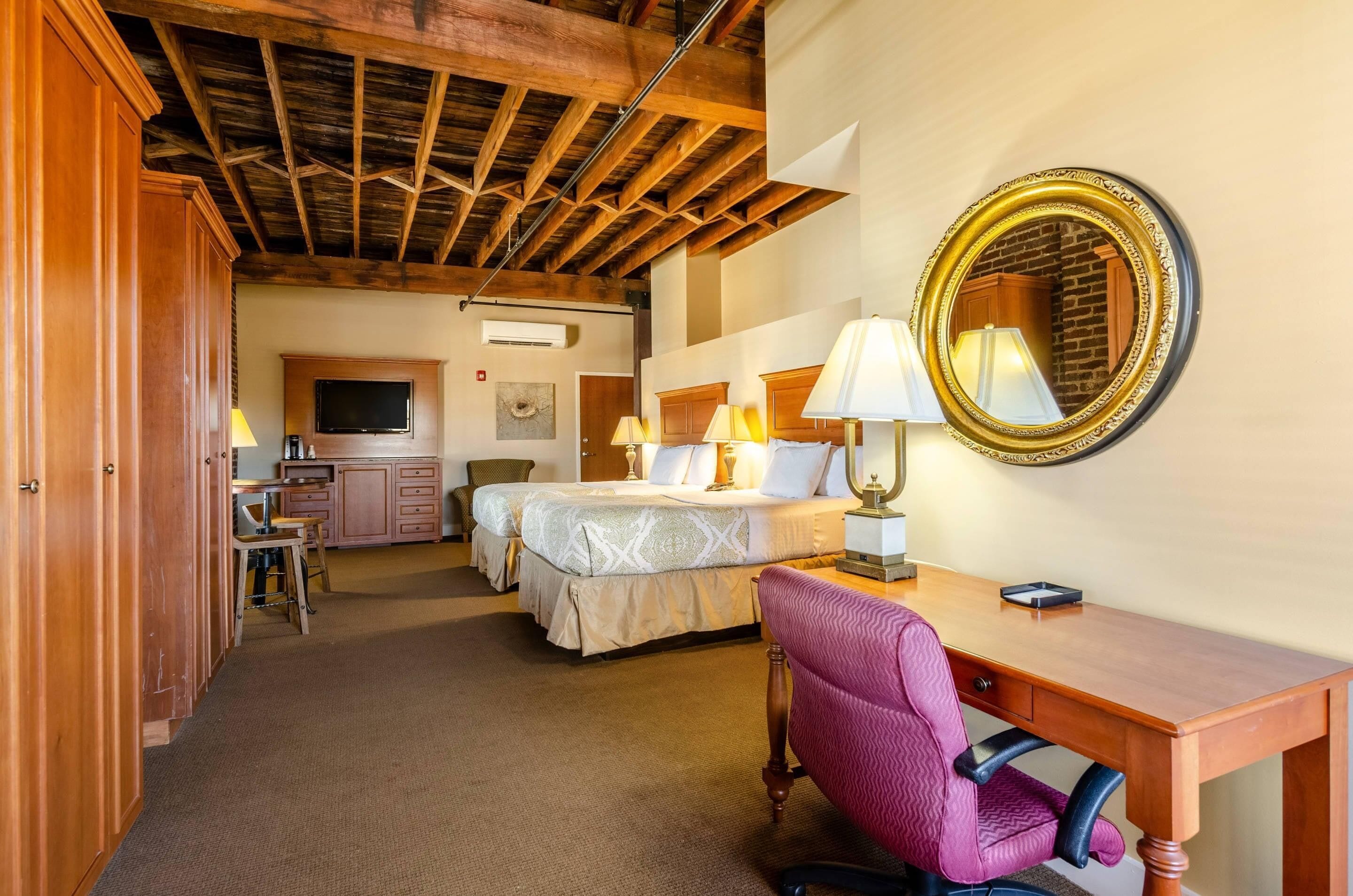 A hotel room with a wooden ceiling, two beds, a desk, a chair, and a mirror.