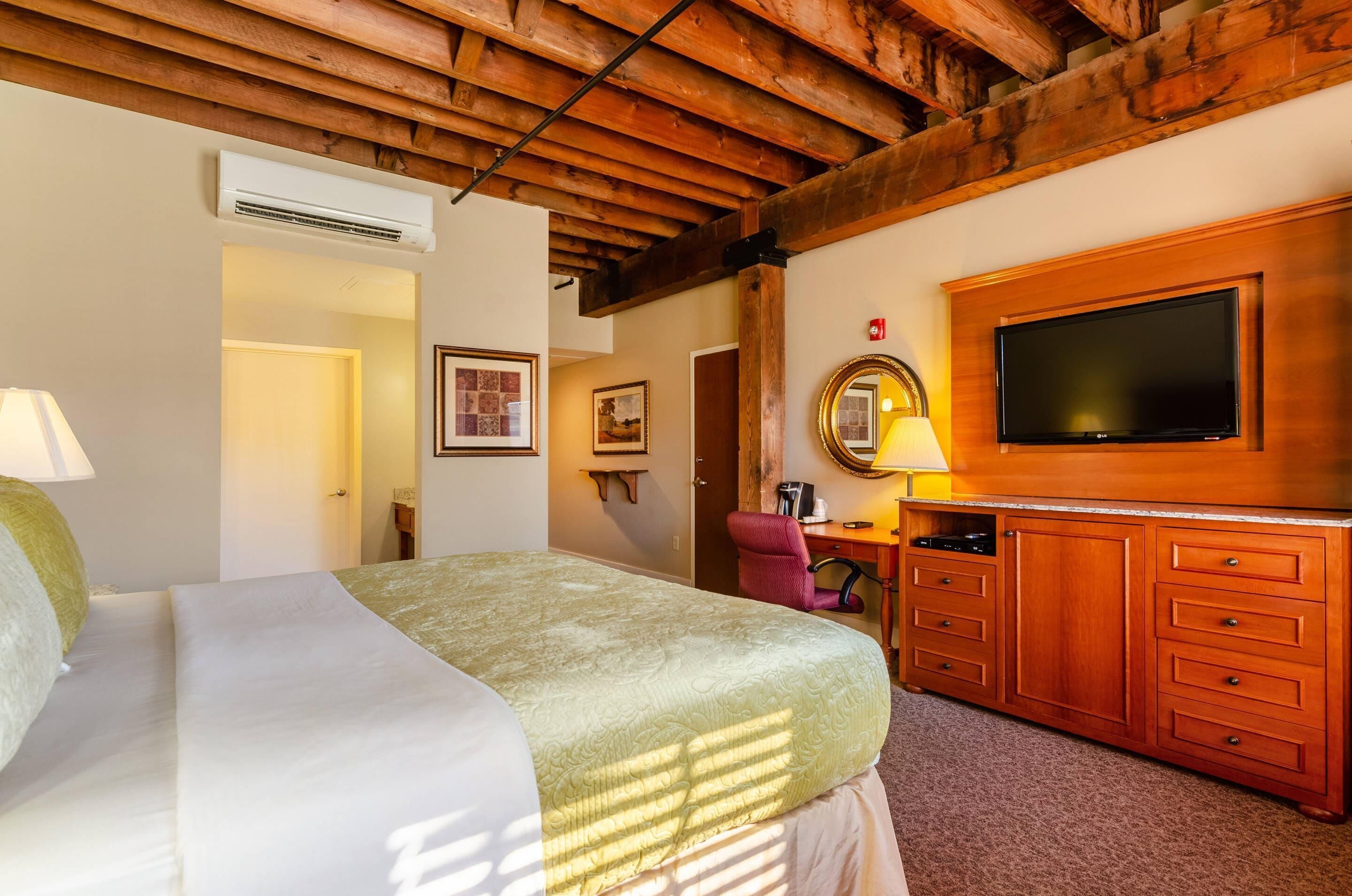 A bedroom with a bed, wooden ceiling beams, a TV, a desk, and a chair.