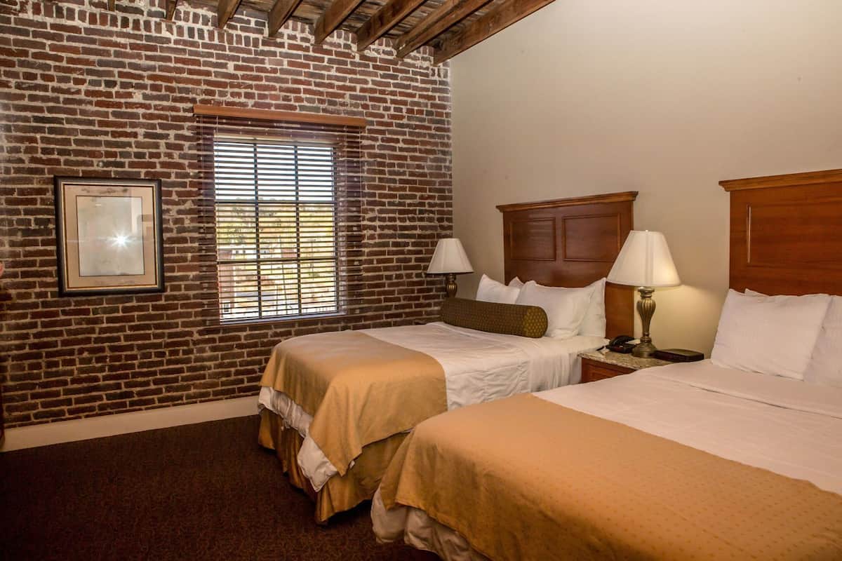 A hotel room with a brick wall, two beds with white linens, a wooden headboard, a window with blinds, and a framed picture on the wall.