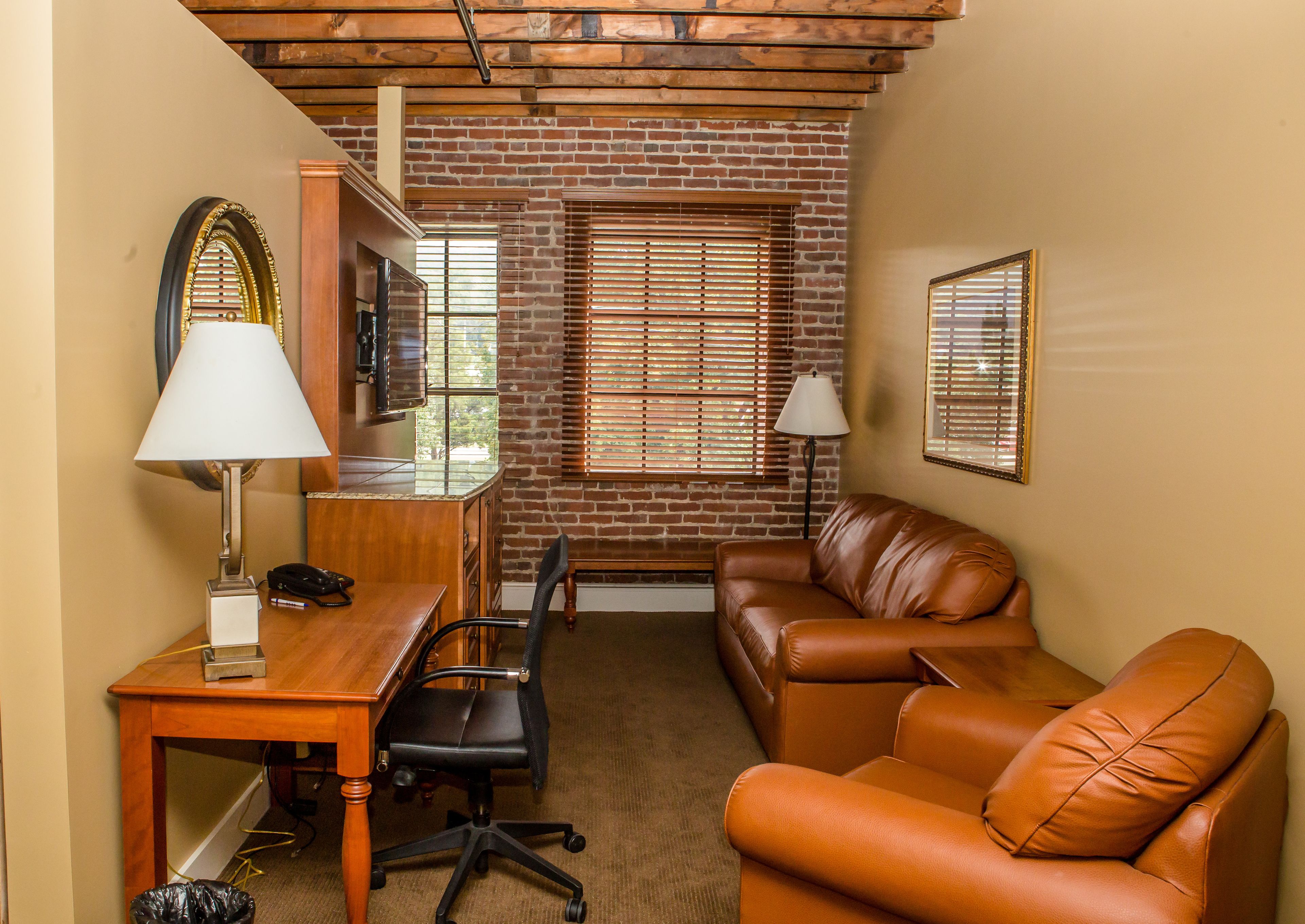 A room with a brick wall, a wooden desk, a leather couch, and two windows with blinds.