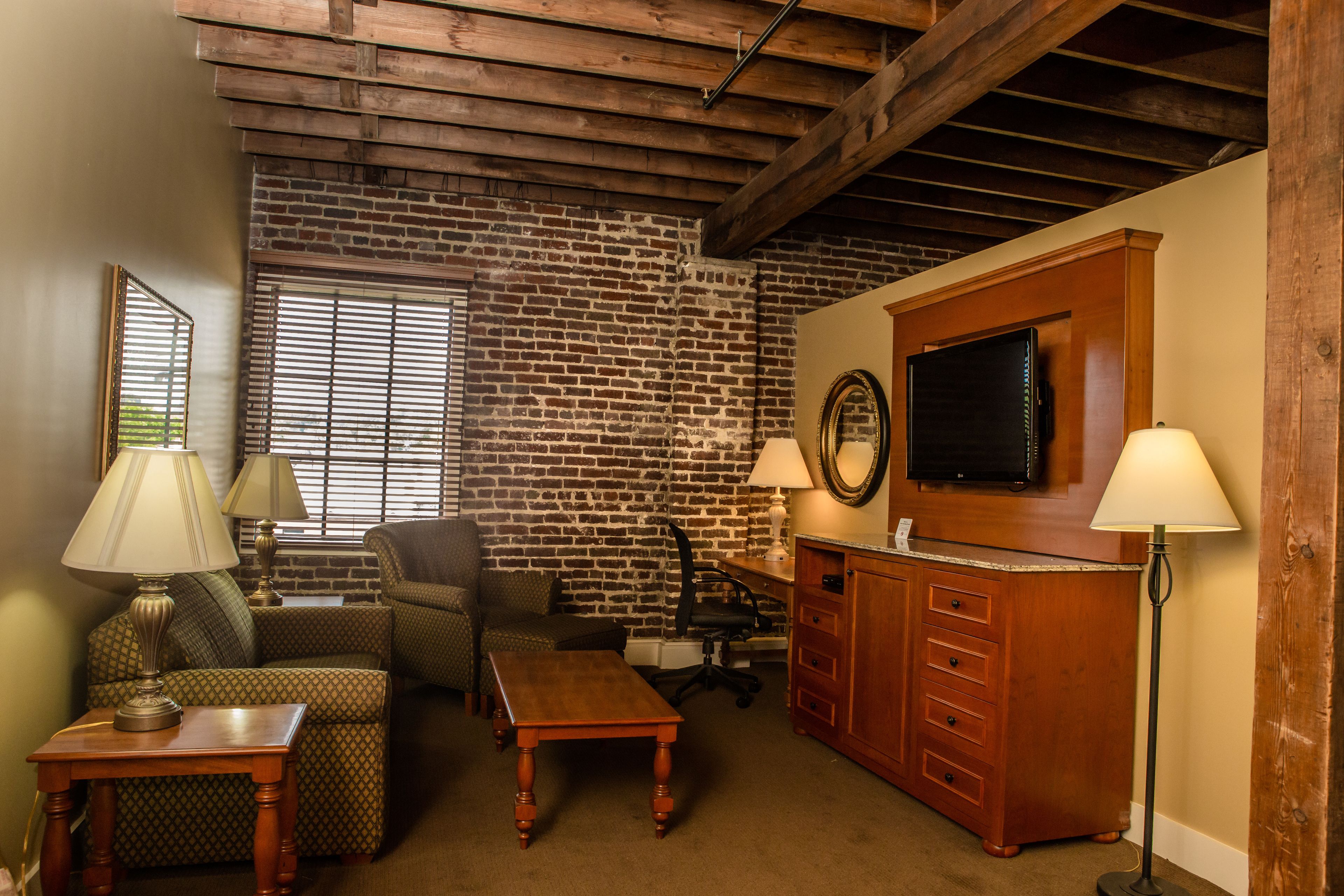 A living room with a brick wall, wooden ceiling beams, a flat-screen TV, a wooden cabinet, and two lamps.