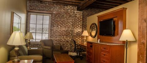A living room with a brick wall, wooden ceiling beams, a flat-screen TV, a wooden cabinet, and two lamps.