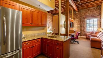 A kitchen with wooden cabinets, a stainless steel refrigerator, granite countertops, and a brick wall.