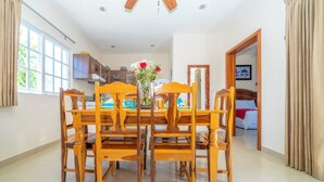 Un comedor con una mesa y sillas de madera, un jarrón con flores y vistas a un dormitorio con cama y cortinas.