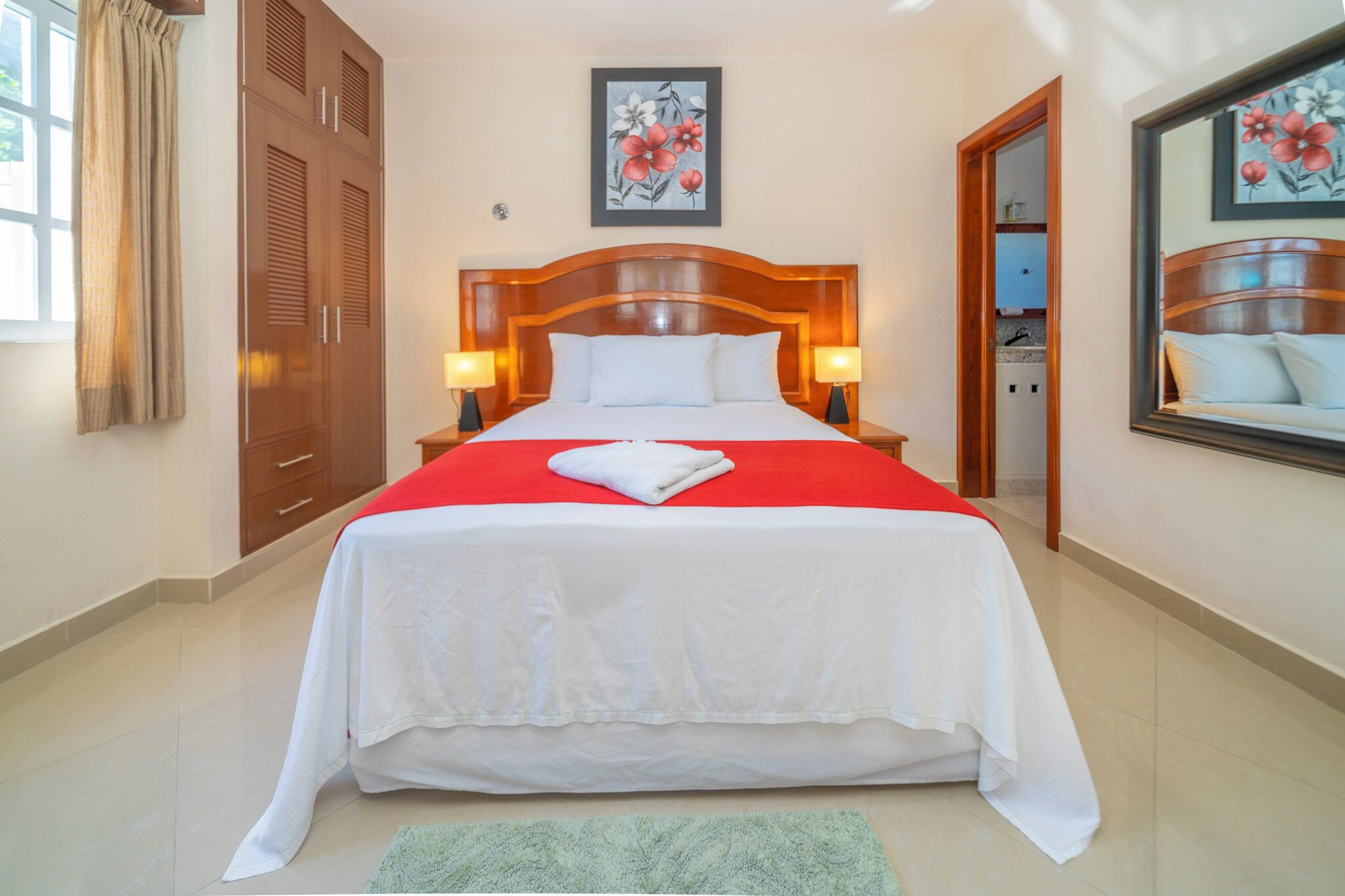 Ein ordentlich bezogenes Bett mit roter Decke und weißen Laken, zwei Nachttischlampen, ein Holz-Kopfteil und ein Blumenbild in einem Rahmen über dem Bett.