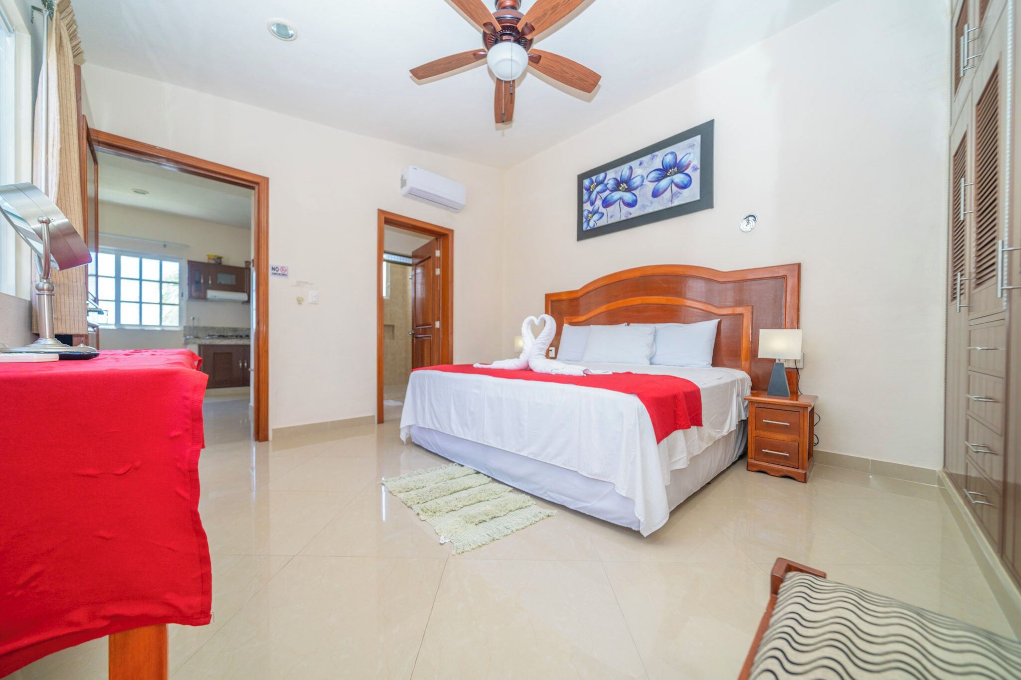 Habitación de hotel con cama, mesita de noche, ventilador de techo, un cuadro y vista a la cocina a través de una puerta abierta.