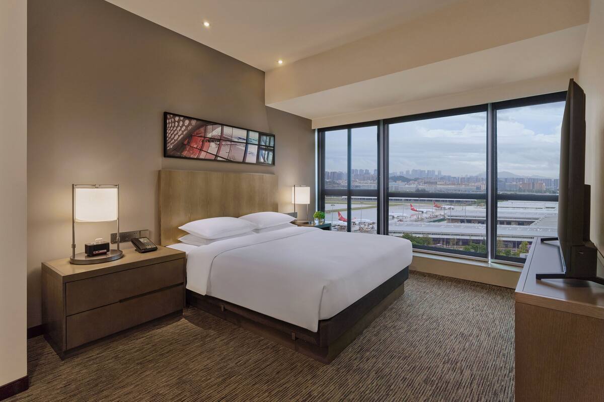 Habitación de hotel con una cama grande, una mesita de noche con lámpara, un escritorio con televisor y un ventanal con vistas a la ciudad.