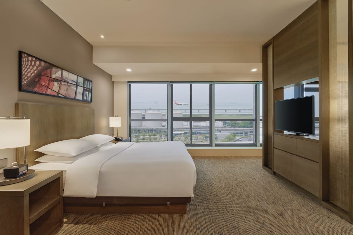 Habitación de hotel con una cama grande, televisión, mesita de noche, lámpara y un ventanal con vistas al aeropuerto y al paisaje urbano.