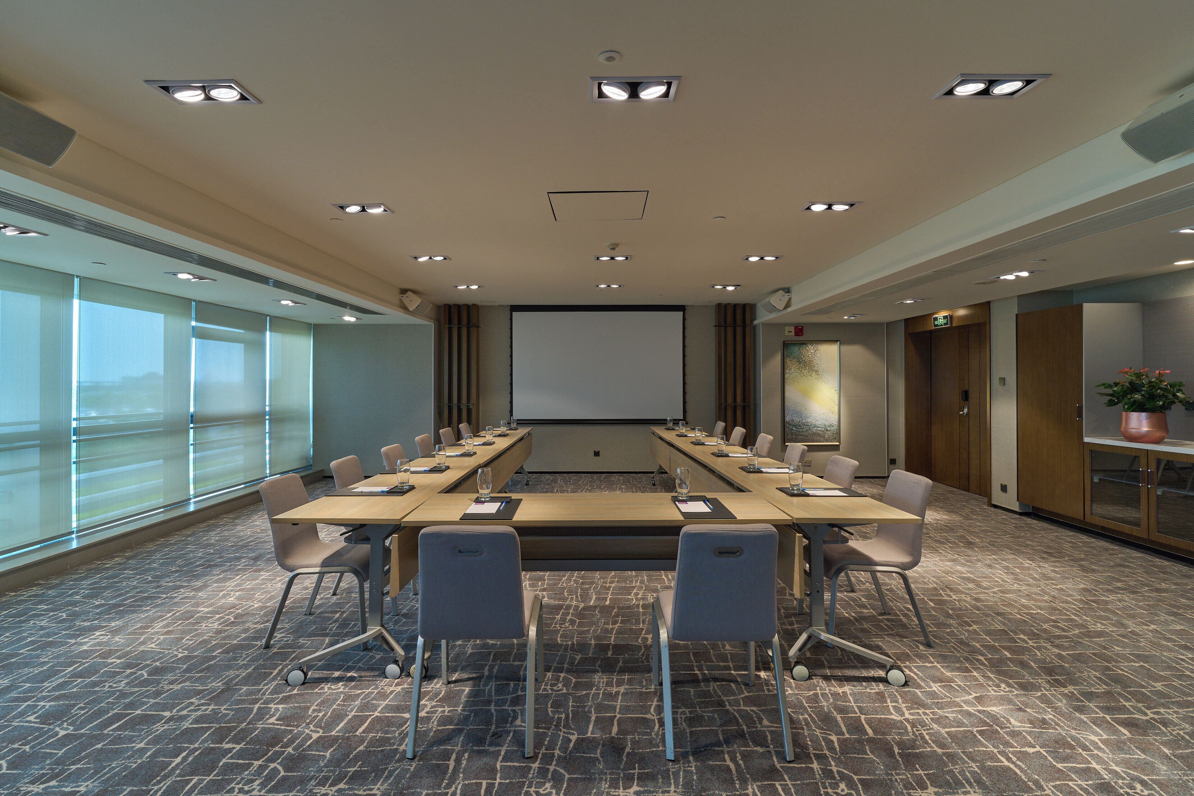 Une salle de conférence équipée d’une longue table, de chaises, d’un écran de projection et de grandes fenêtres.