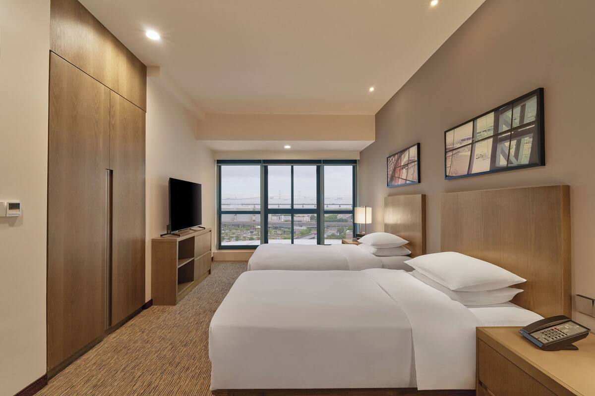 Habitación de hotel con dos camas, televisión, escritorio con teléfono y ventanas amplias que dan a vistas de la ciudad.