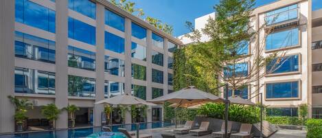 A modern hotel courtyard with a swimming pool, lounge chairs, and large umbrellas.