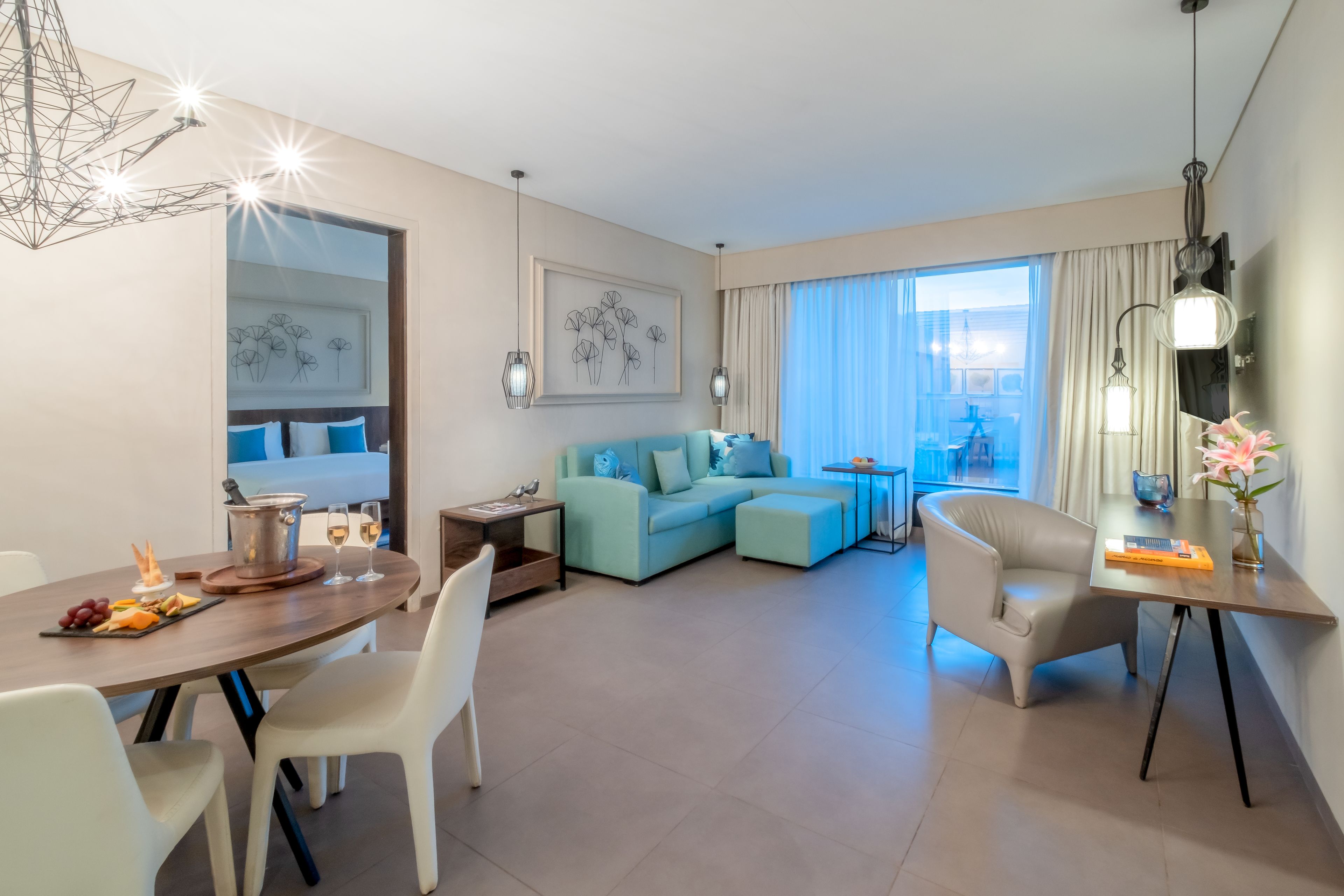 Una habitación de hotel moderna con zona de comedor, un sofá azul claro, una mesa redonda con sillas blancas y un ventanal con cortinas.