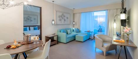 Una habitación de hotel moderna con zona de comedor, un sofá azul claro, una mesa redonda con sillas blancas y un ventanal con cortinas.