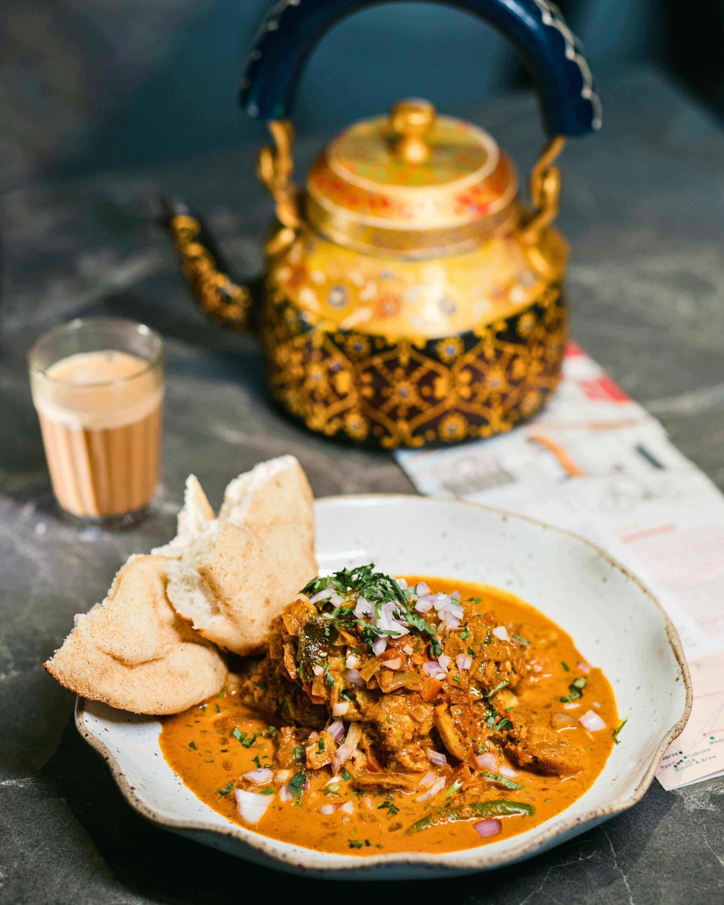 Un plato de curry con pan y un vaso de té, una tetera decorativa y un menú sobre una mesa.