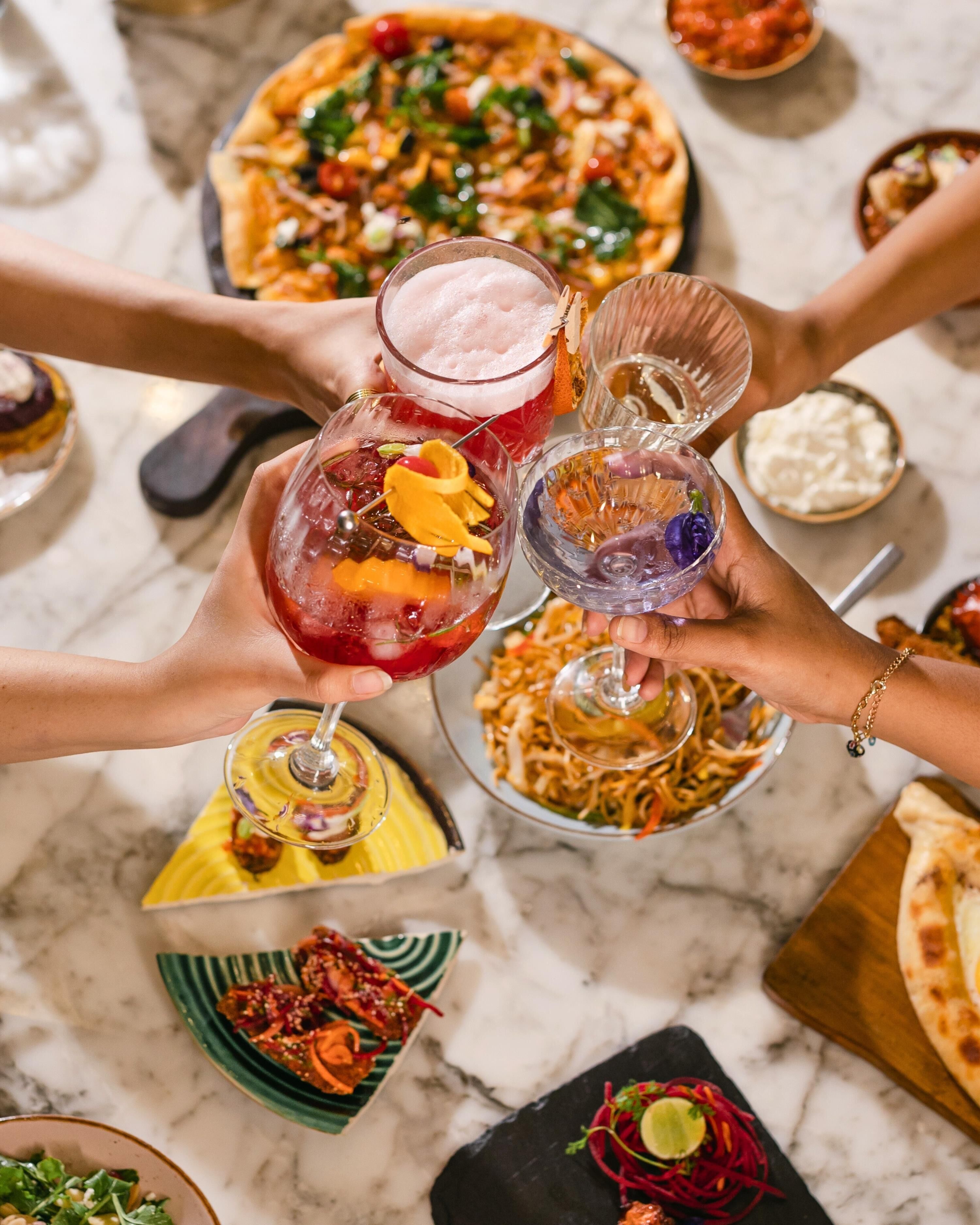 A group of people holding drinks at a table with various dishes and a pizza.