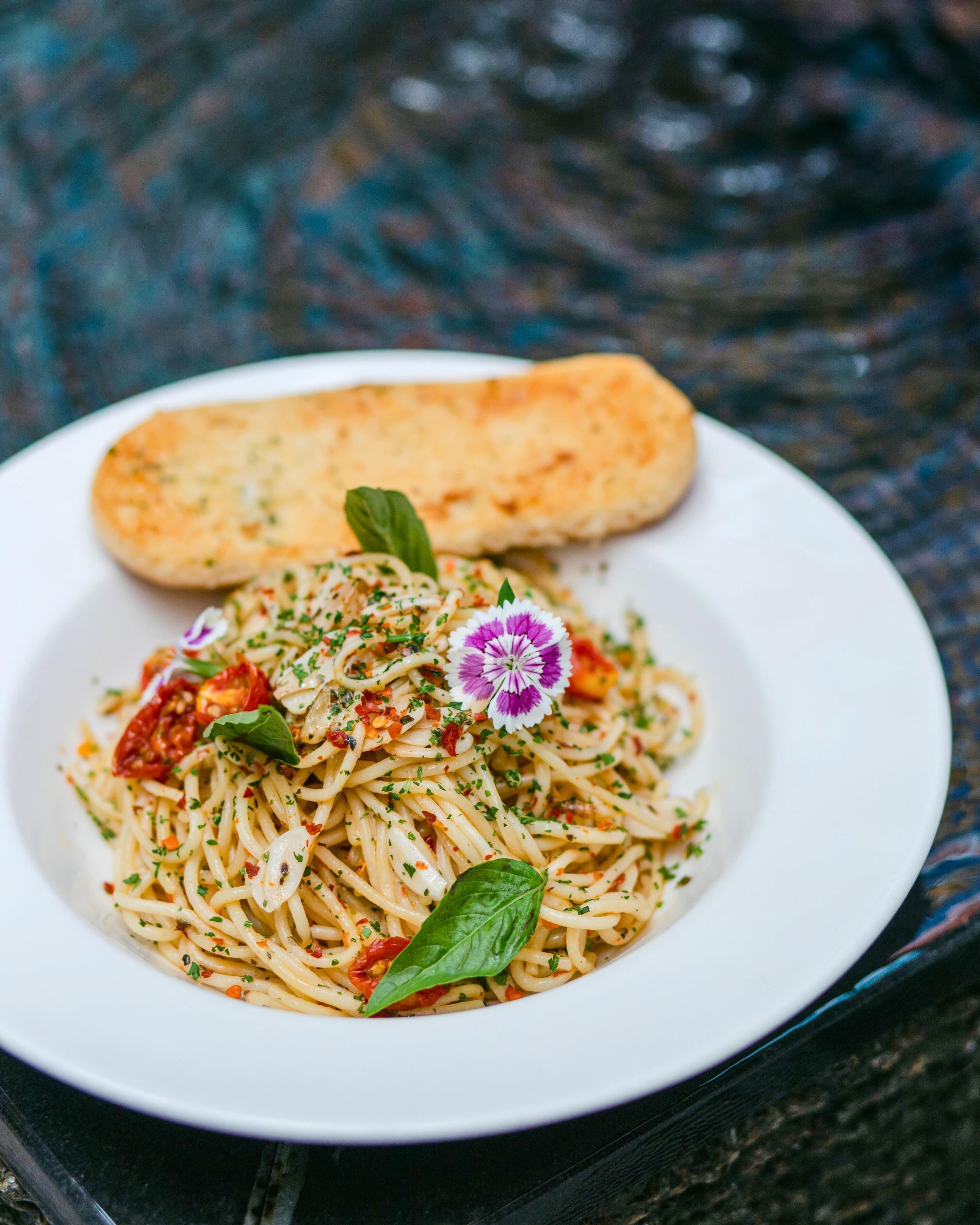A plate of spaghetti with a basil leaf garnish, a small purple flower, and a piece of bread on a white plate.