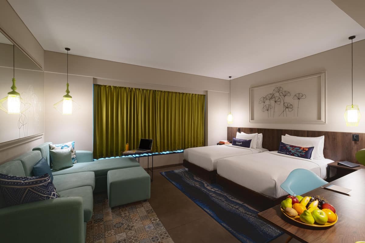 Quarto de hotel com cama, escrivaninha, cadeira, sofá, laptop na escrivaninha e uma fruteira sobre a mesa.