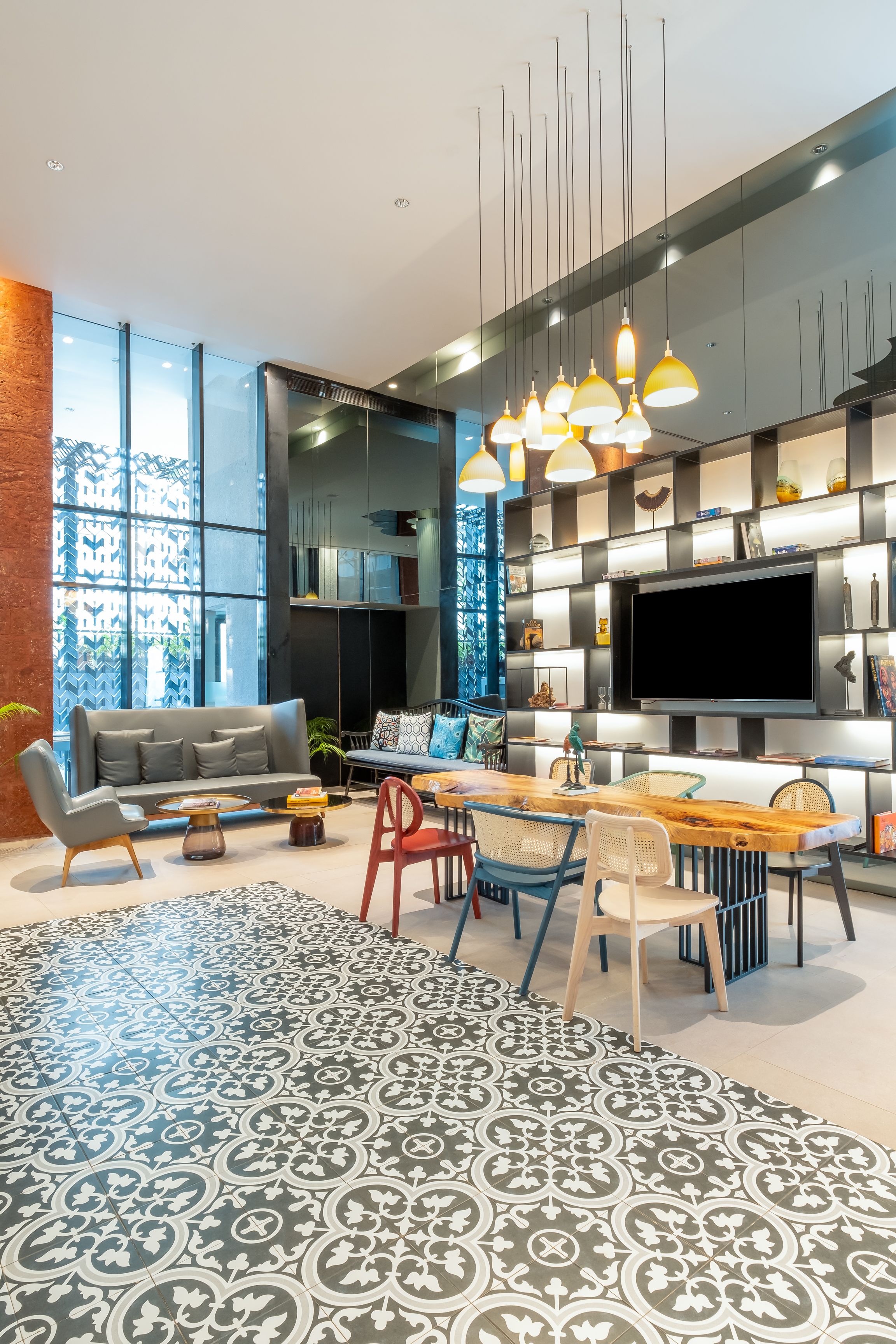 A modern living room with a sofa, coffee table, dining table, and chairs. There is a large TV mounted on the wall, and a bookshelf with various items.