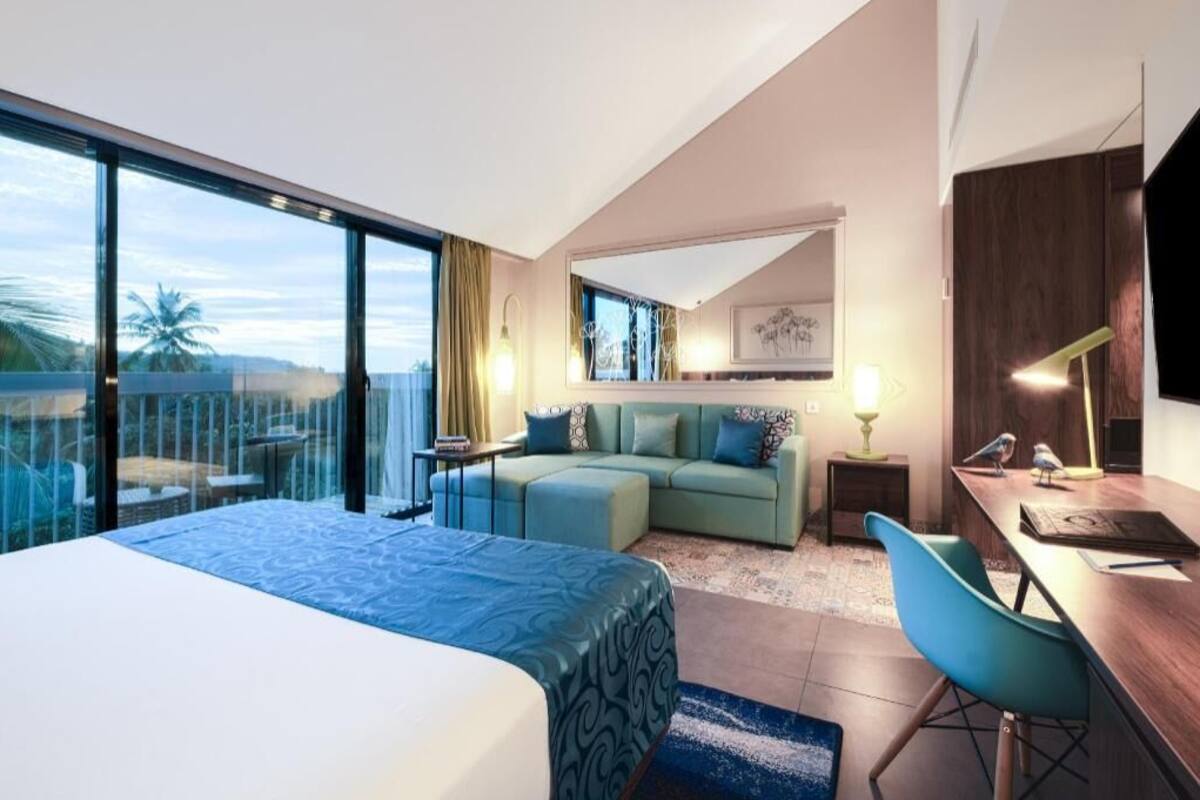 Quarto de hotel moderno com uma cama grande, uma escrivaninha com cadeira, um sofá e uma varanda com vista.