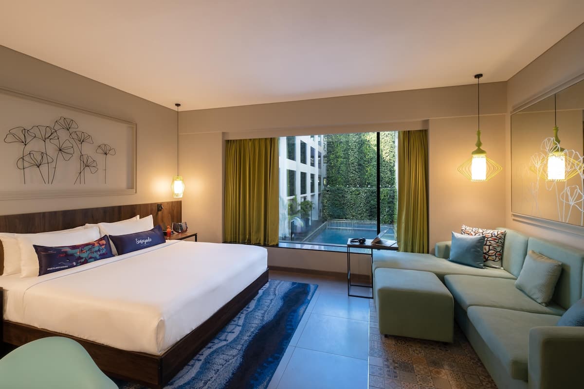 Quarto de hotel moderno com uma cama grande, um sofá, uma divisória de vidro e vista para uma piscina e edifícios.