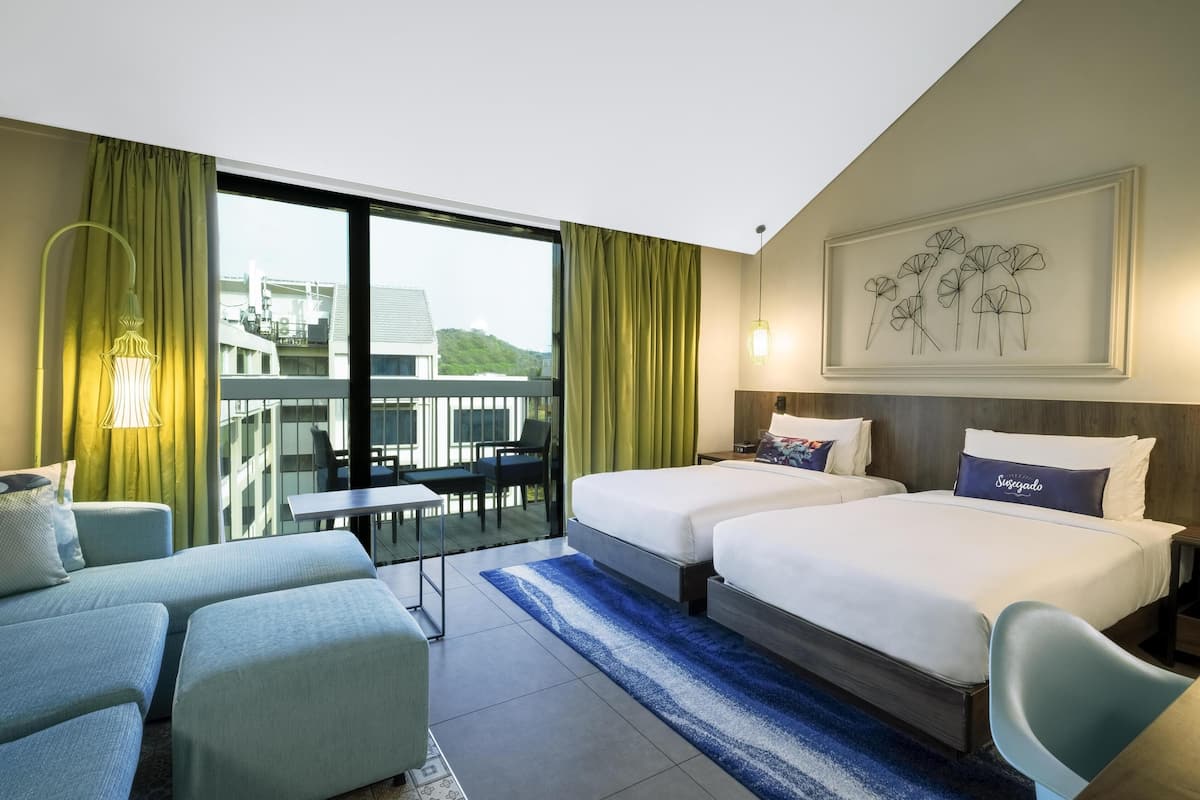 Quarto de hotel com duas camas, varanda com mesa e cadeiras, e uma grande janela com cortinas.