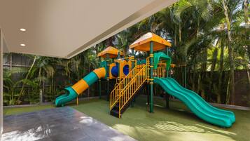A children's play area with slides and climbing structures, surrounded by palm trees.