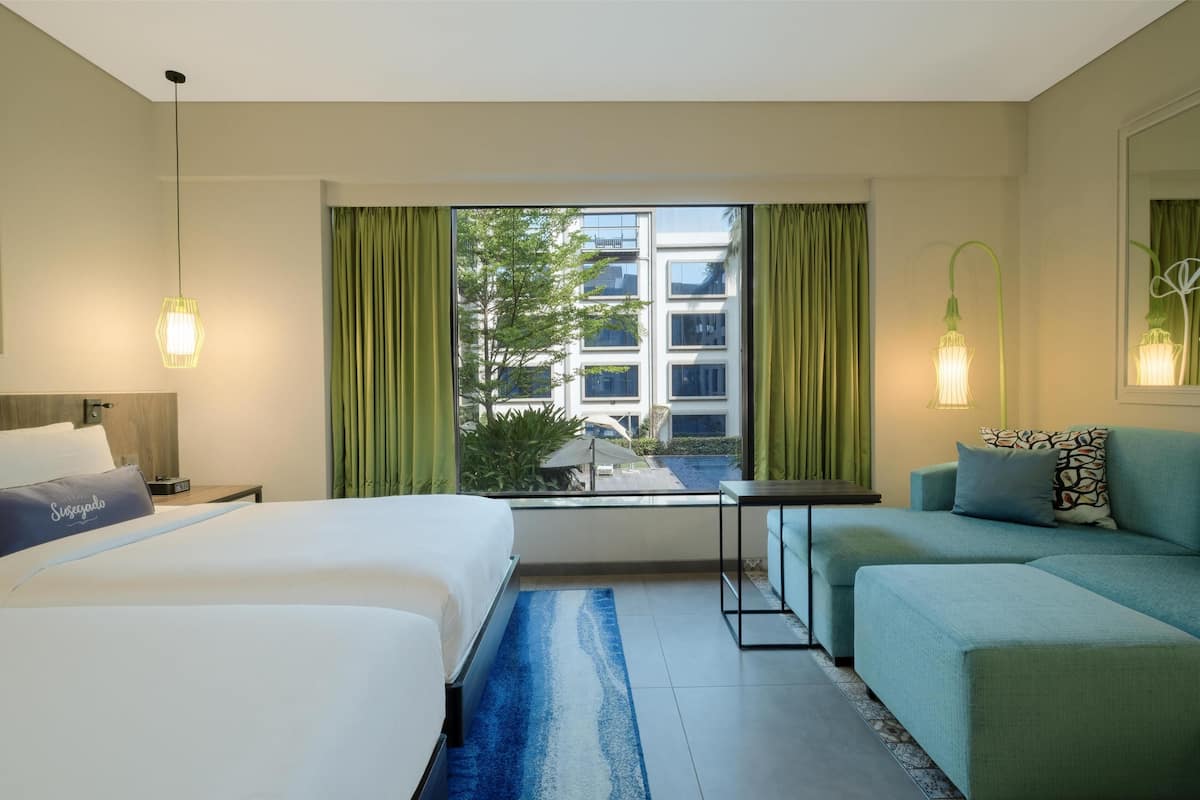 Quarto de hotel com cama, sofá, mesinha e janela com cortinas.