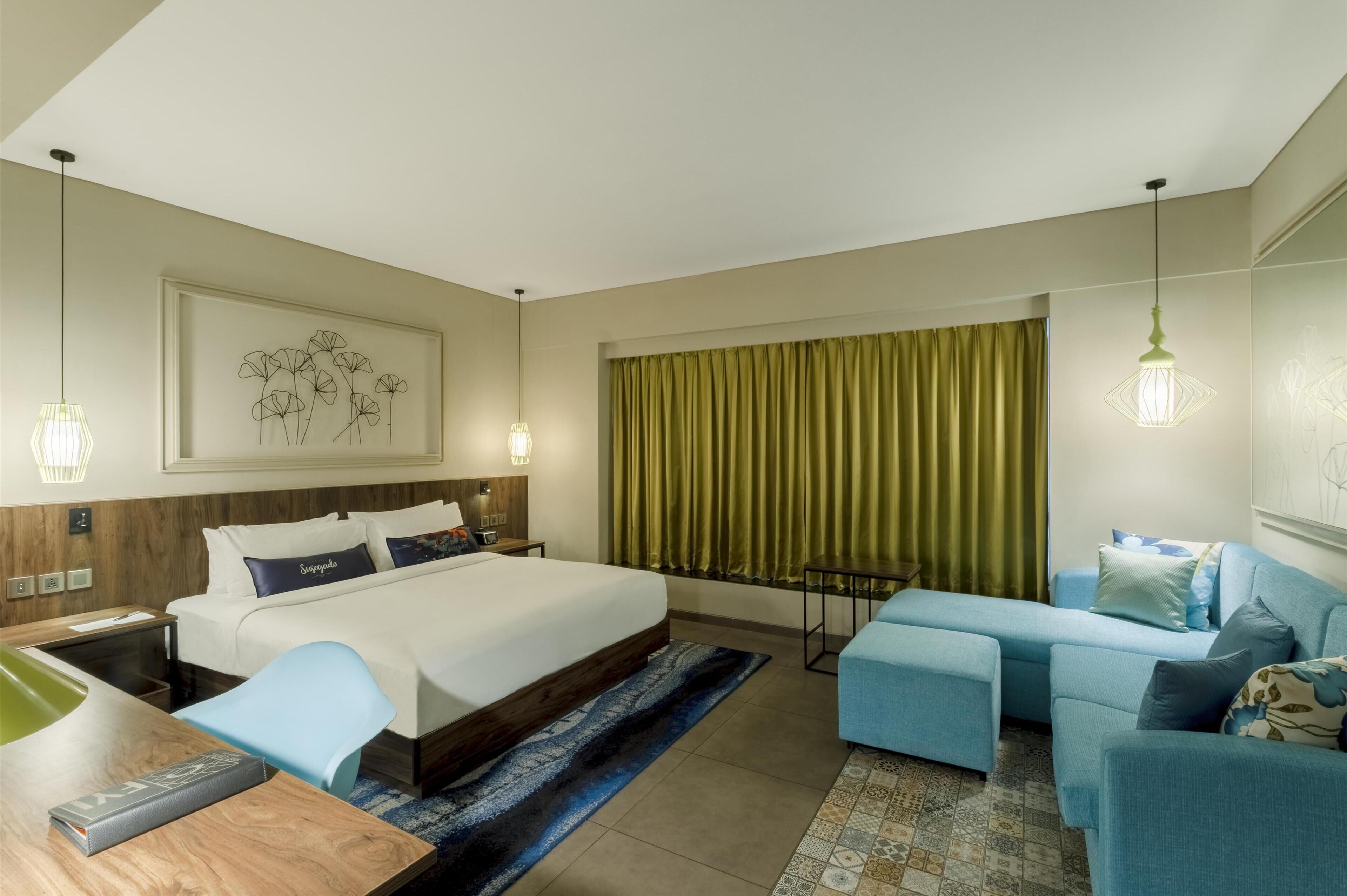 A hotel room with a large bed, a desk with a chair, a sofa, and a window with curtains.