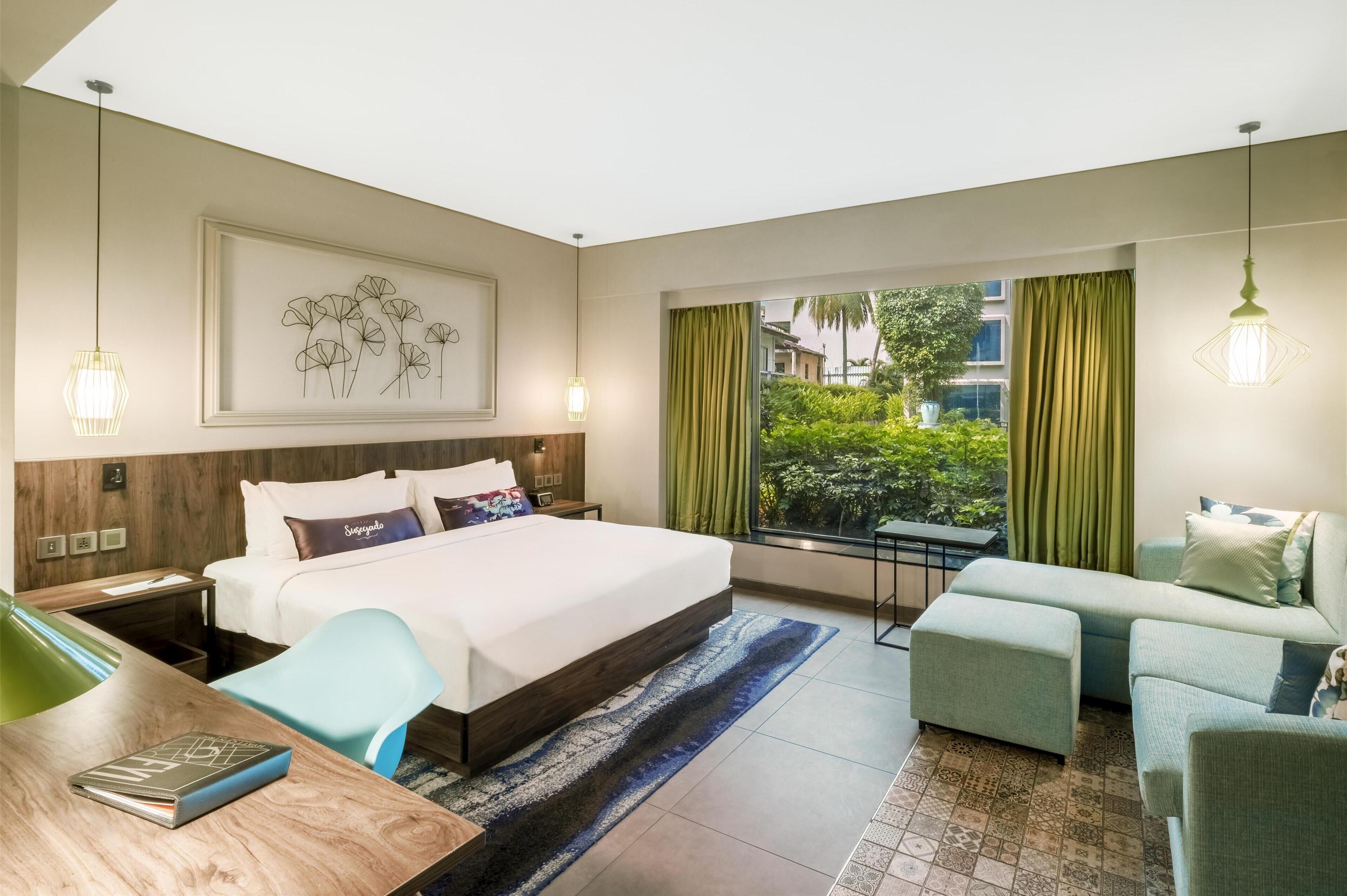 A hotel room with a large bed, a desk, a sofa, and a view of greenery outside.