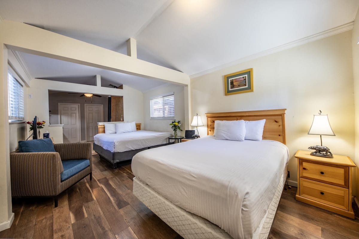 Habitación de hotel con dos camas, un sofá, suelo de madera y una mesita de noche con lámpara.