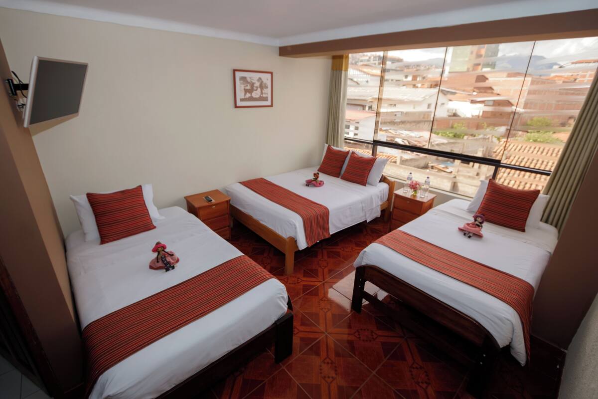 A hotel room with three beds, a TV, a framed picture on the wall, and a view of the city through large windows.
