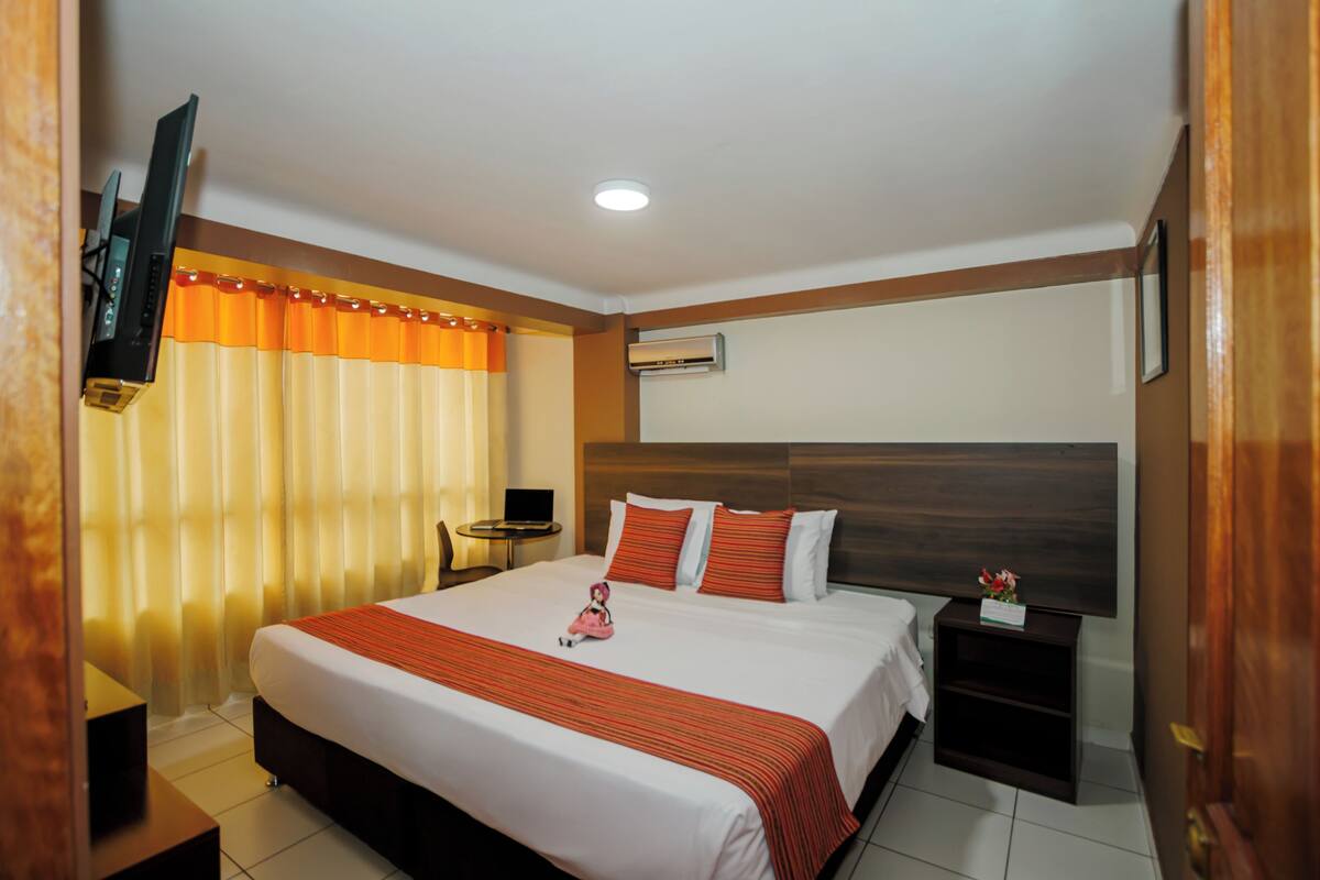 A hotel room with a large bed, a TV, a small table, and an air conditioning unit.