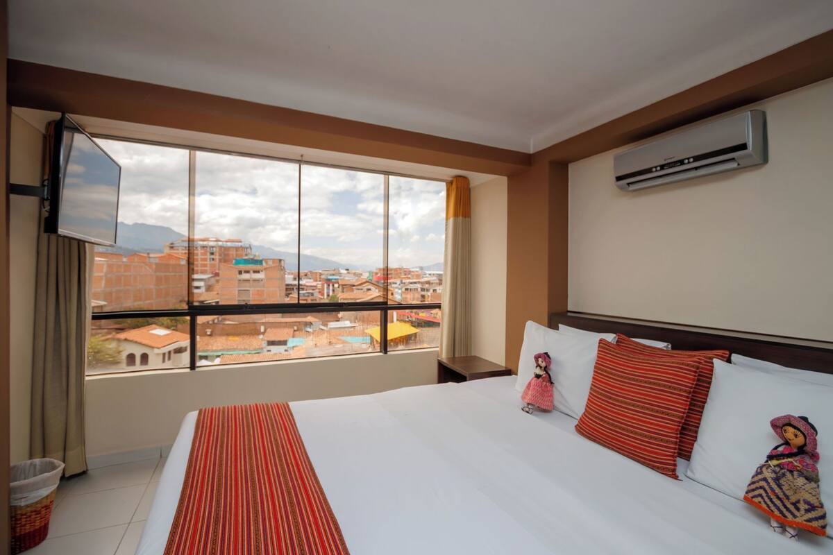 A hotel room with a large bed, a flat-screen TV, an air conditioning unit, and a view of the cityscape.