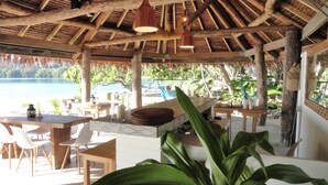 An outdoor seating area with wooden tables and chairs, a thatched roof, and a view of the sea. - Two Canoes Island Getaway (Aore Island)
