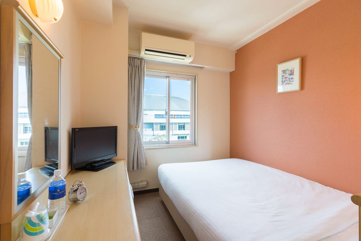 Quarto de hotel com uma cama, televisão, escrivaninha com uma garrafa de água, relógio e janela com cortina.