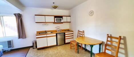 A compact kitchen with a microwave, sink, and small refrigerator. There is a wooden table with chairs and a clock on the wall.