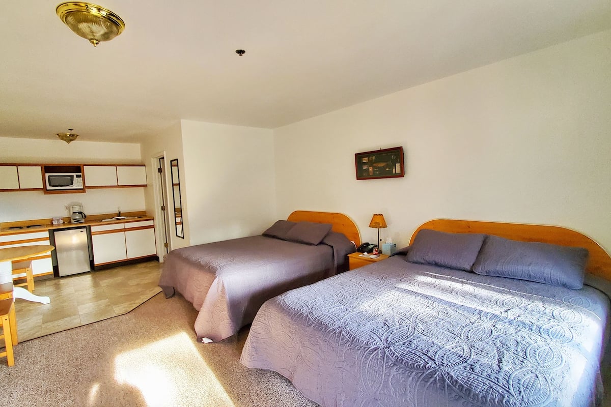 A compact hotel room with two beds, a kitchenette, a small dining area, and a bathroom.