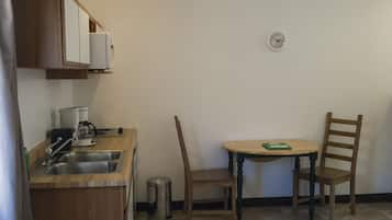 Cocina pequeña con un lavamanos, una cafetera y un bote de basura. Hay una mesa de madera con dos sillas y un reloj en la pared.