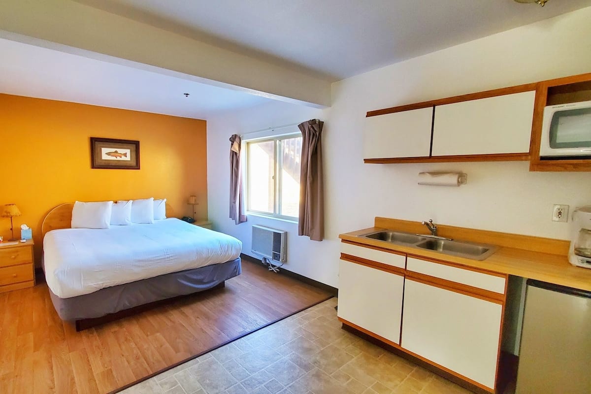 A hotel room with a bed, a small bedside table, a lamp, a framed picture on the wall, a window with curtains, a kitchenette with a sink and cabinets, and a microwave above the counter.