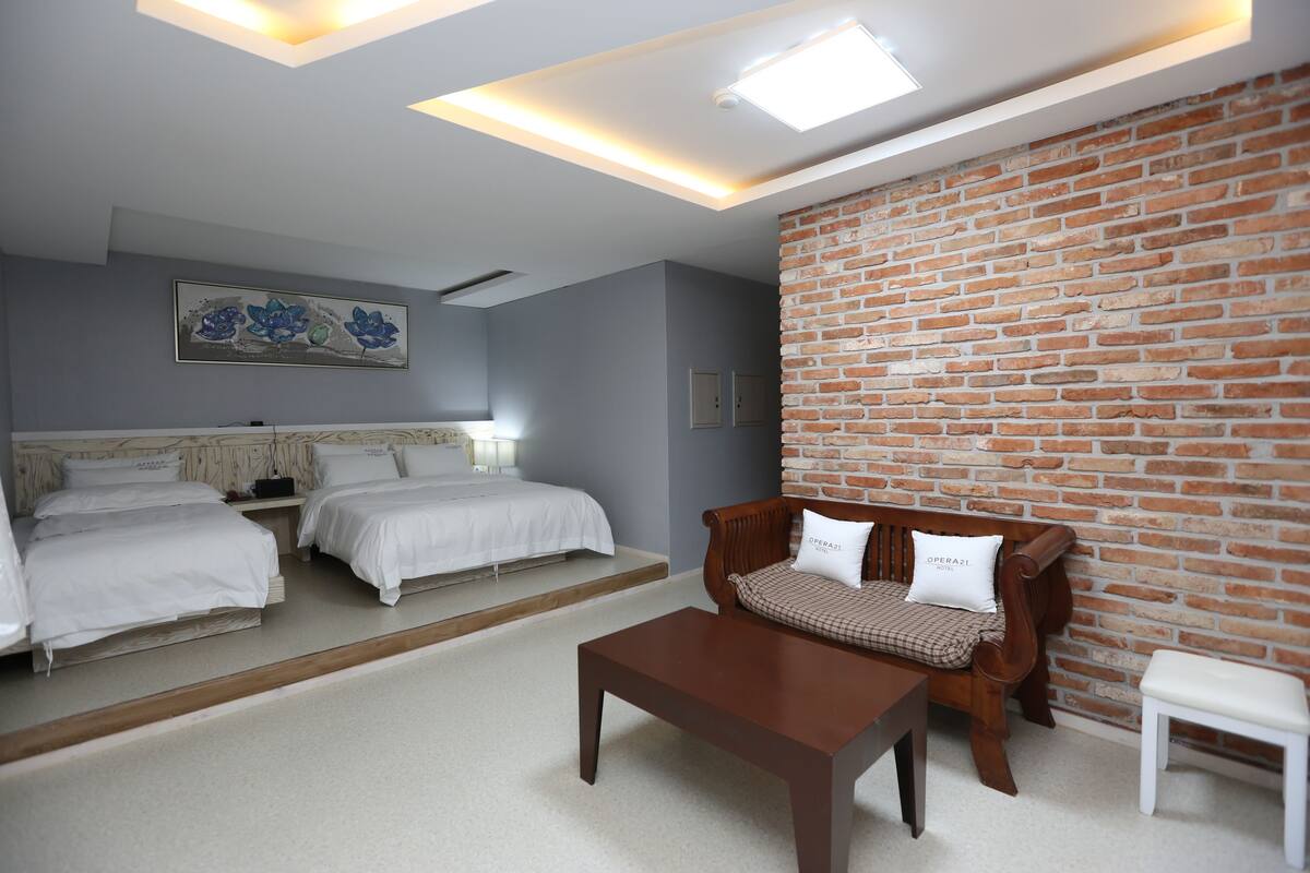 Ein Hotelzimmer mit zwei Betten, einer Holzbank, einem kleinen Tisch und einer Ziegelwand.