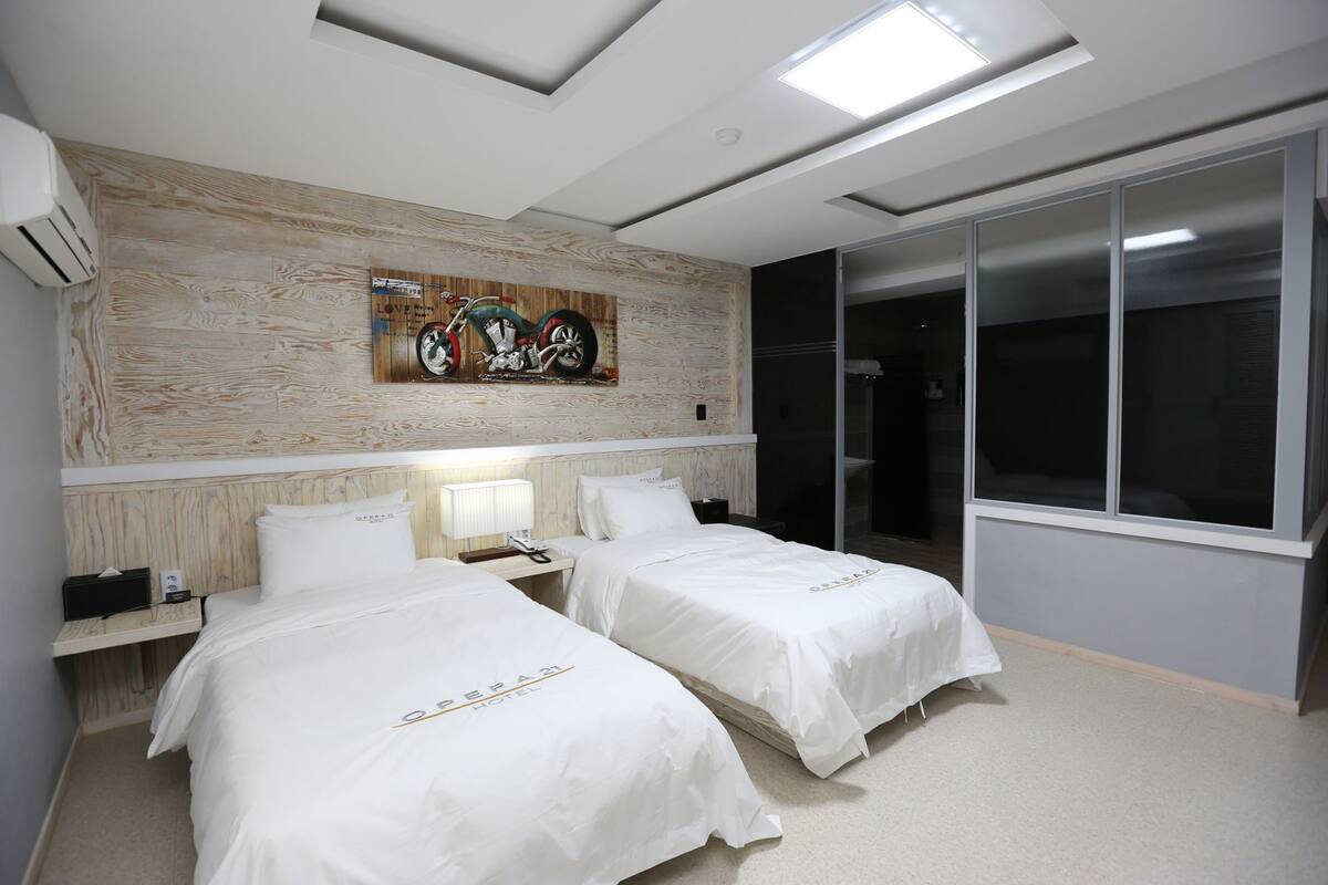 Ein Hotelzimmer mit zwei Betten, einem Holzkopfende, einem an der Wand hängenden Kunstwerk, einem Nachttisch mit Lampe und einer Schiebetür zu einem Badezimmer.