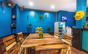 A dining area with a wooden table and chairs, a refrigerator, and hanging plants. - BGW Phuket - Hostel (Patong)