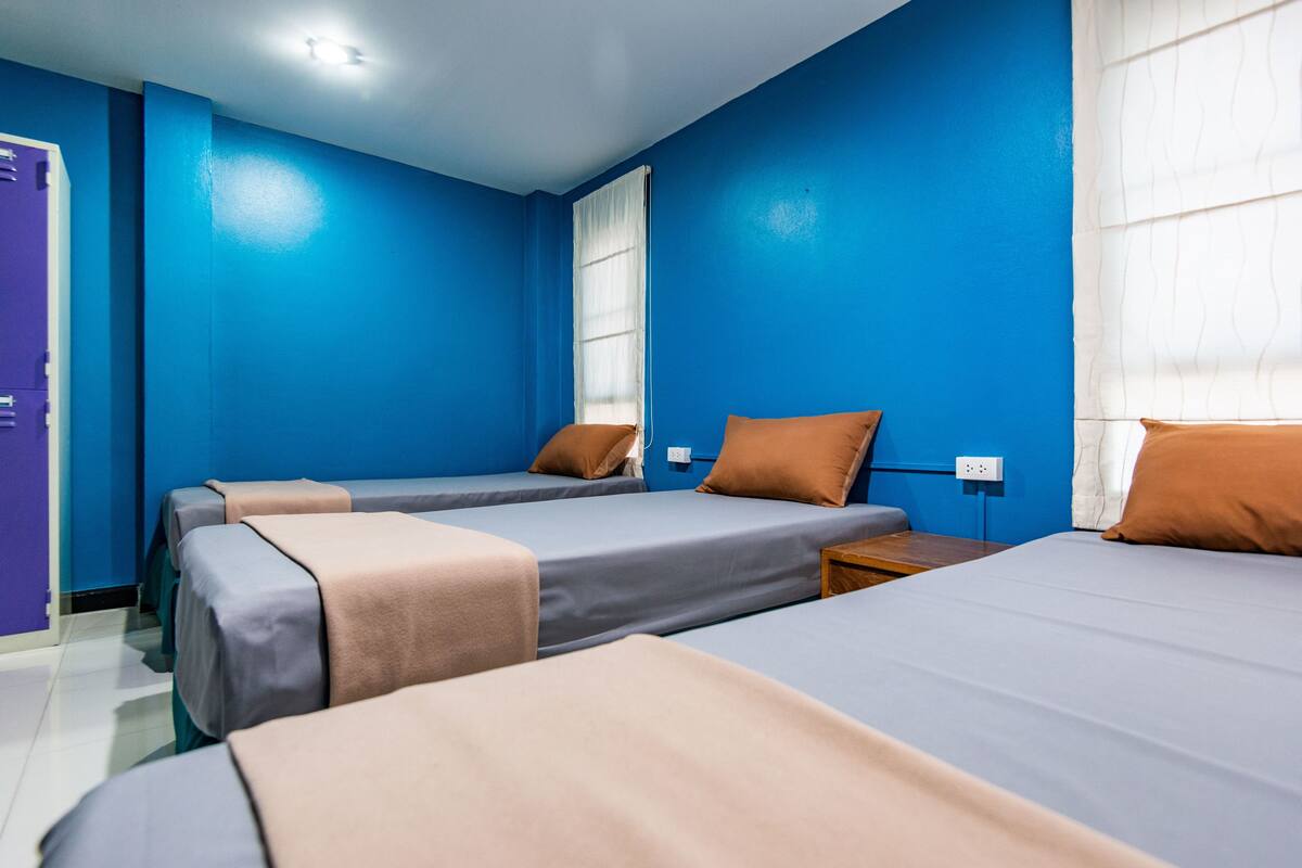 A hotel room with three beds, each with a different colored bedspread, and a window with white curtains.