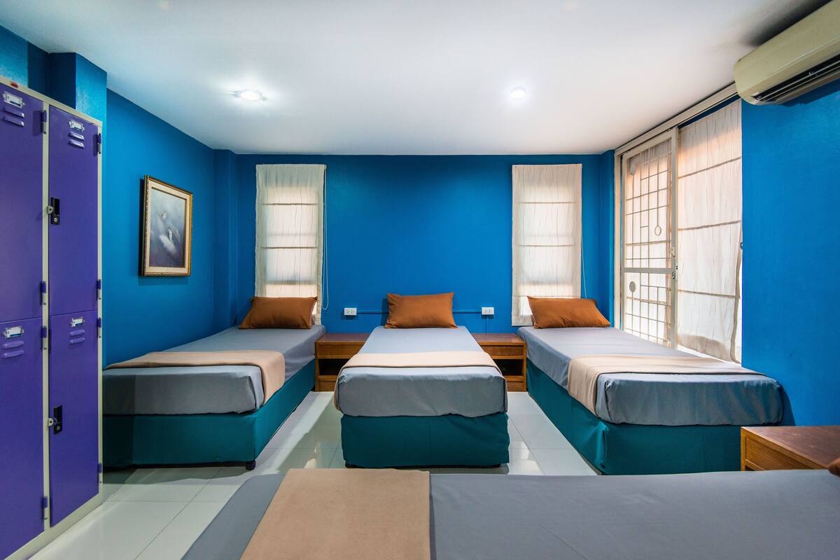 A room with three single beds, a wooden nightstand between each bed, and a large window with blinds.