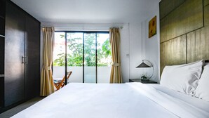 A hotel room with a large bed, a chair, a nightstand with a lamp, and a view of greenery through the window. - BGW Phuket - Hostel (Patong)