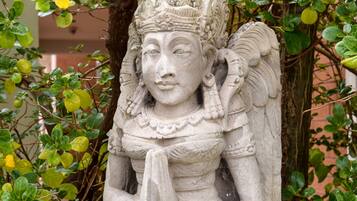 A stone statue of a figure with a crown, wings, and a floral pattern on the garment, set against a backdrop of green foliage and a building.