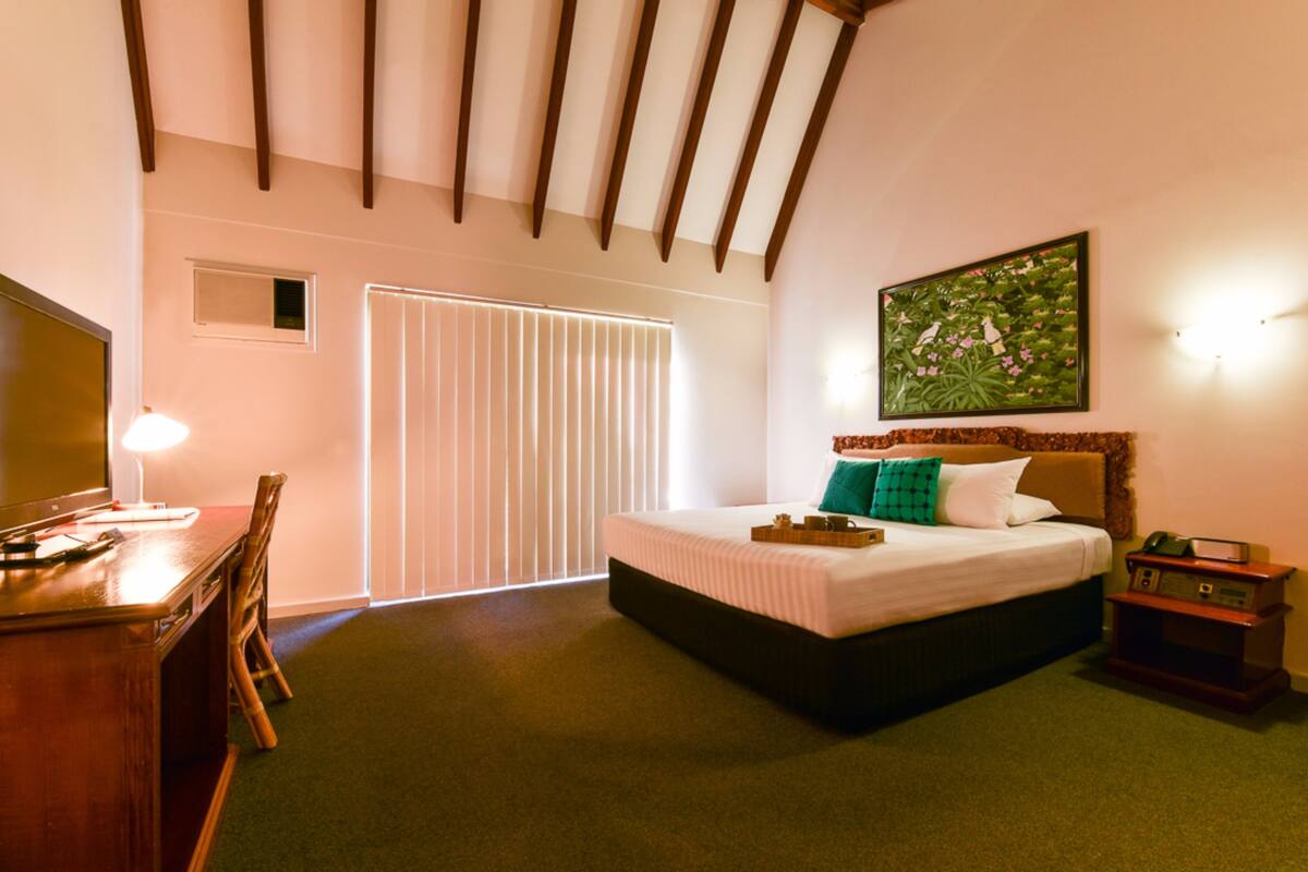 A hotel room with a bed, a desk, a chair, a TV, a painting on the wall, and a window with blinds.