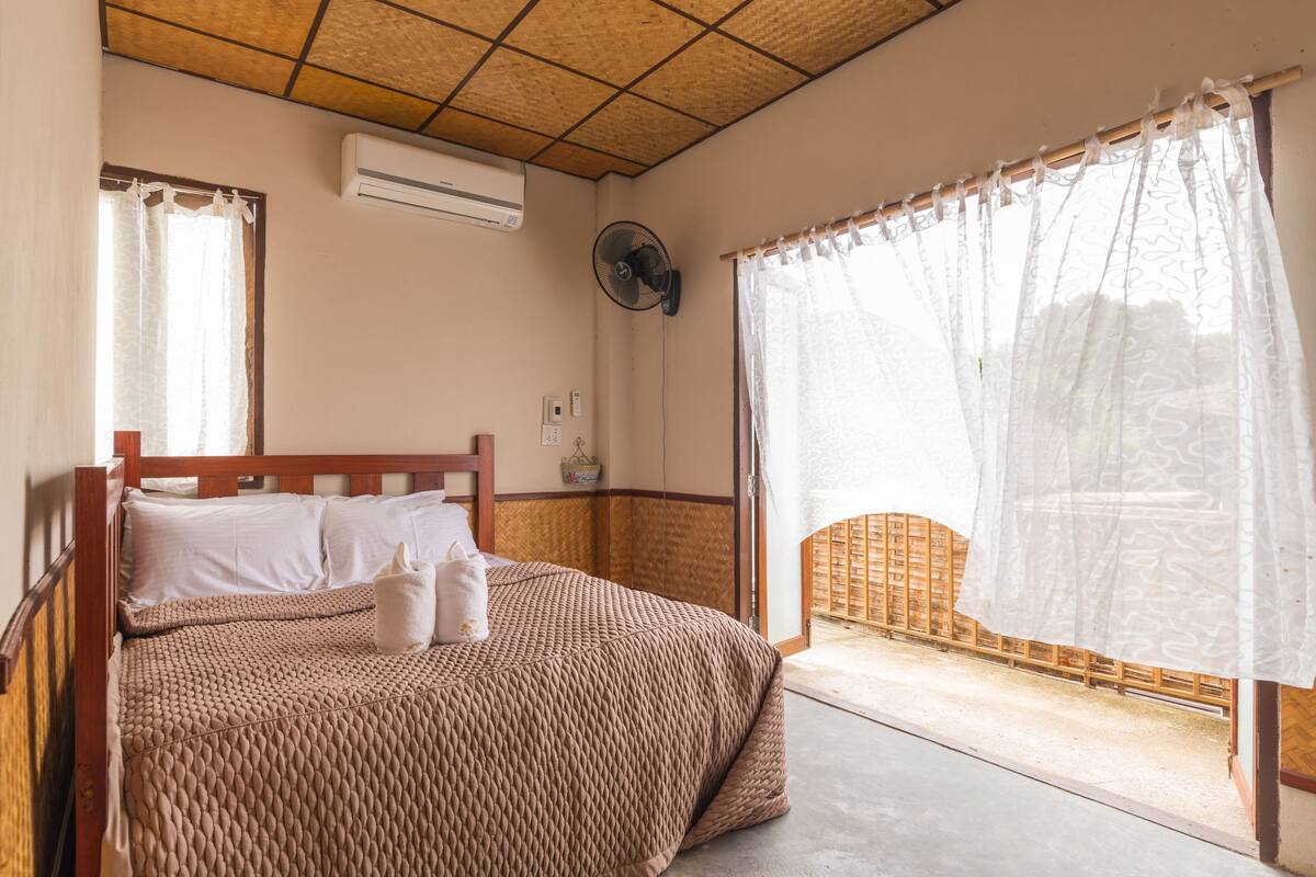 Un dormitorio con cama, ventilador de techo y ventana con cortinas translúcidas.