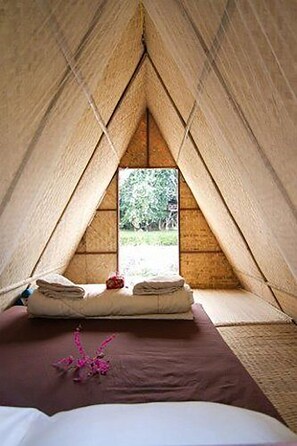 Quarto aconchegante em formato triangular, com cama, almofadas e janela com vista para o verde.