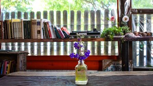 A wooden table with a clear glass vase containing purple flowers, a bookshelf with books, and a view of a wooden fence and trees through a window. - B&B Hostel (Punta del Este)